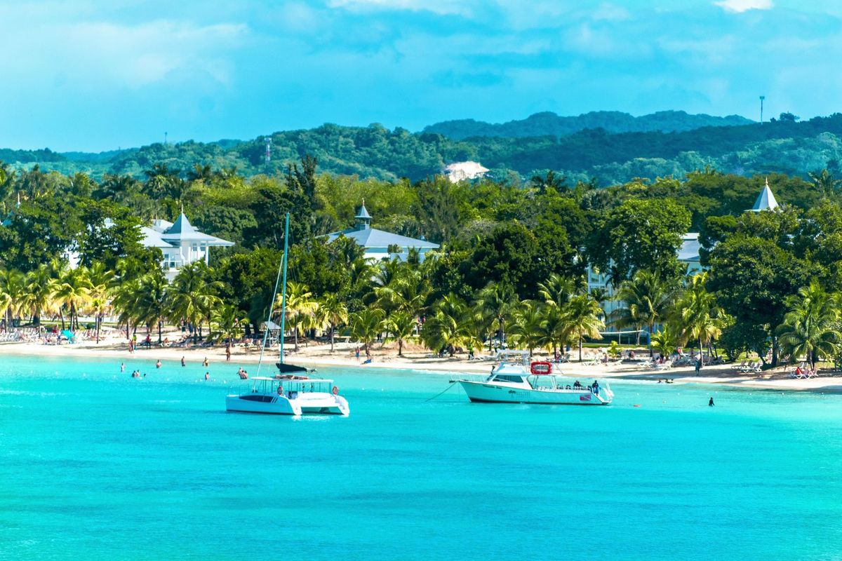 A turquoise bay in Jamaica with a catamaran and a beach lined with palm trees and resort buildings